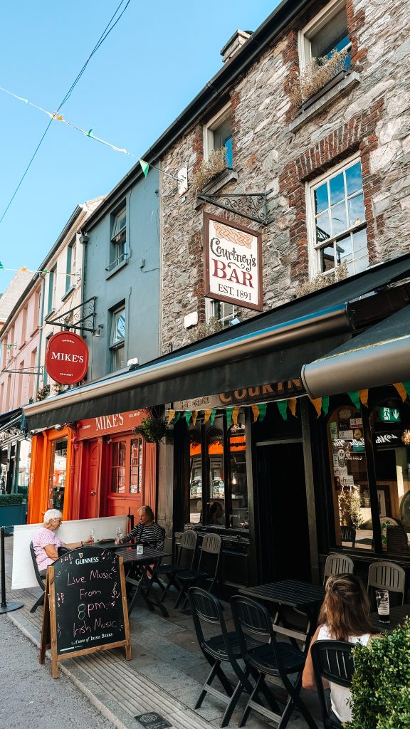 A stone grey pub with a black awning next to a cobblestone street with tables and people sitting underneath the awning.