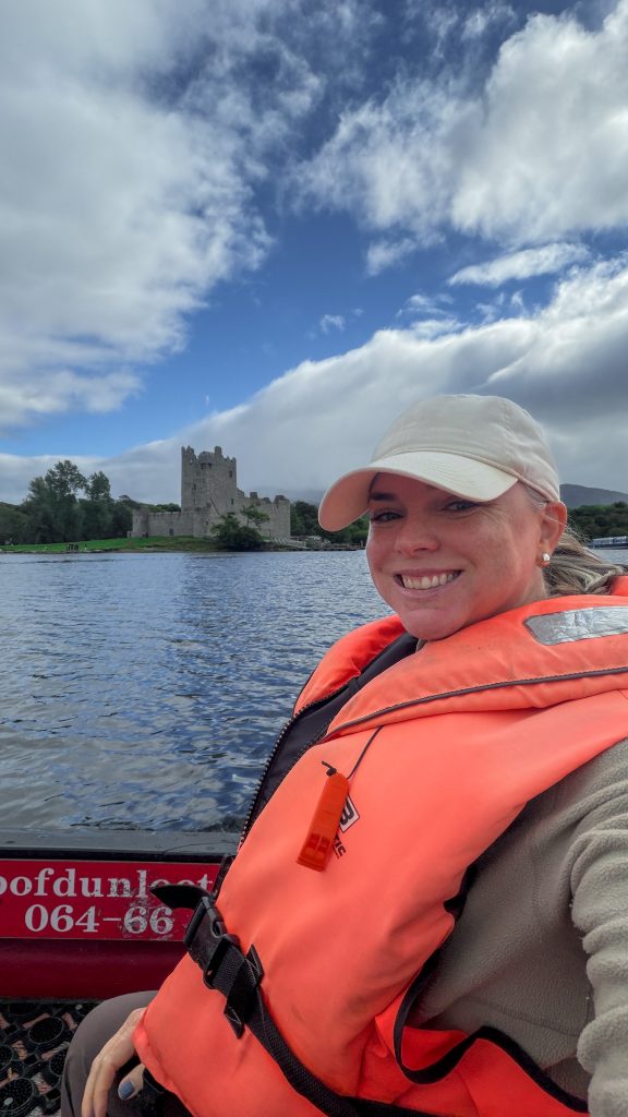 Woman on a boat in a lake wearing an orange life vest and white hat smiles at the camera with a stone castle in the background.