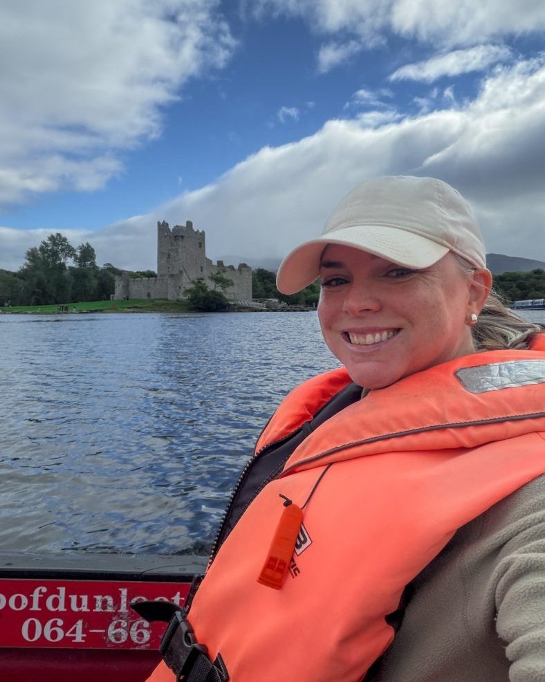 Woman on a boat in a lake wearing an orange life vest and white hat smiles at the camera with a stone castle in the background.