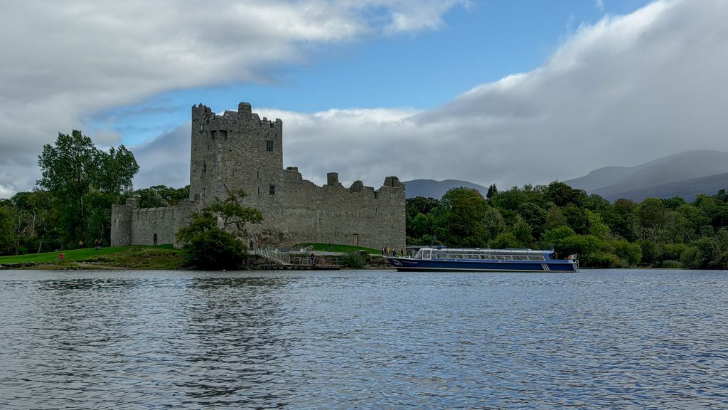 Stone castle seen from a lake with a cloudy sky and mountains in the distance.