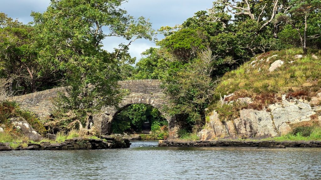 An old stone arch bridge crosses a lake surrounded by green vegetation.