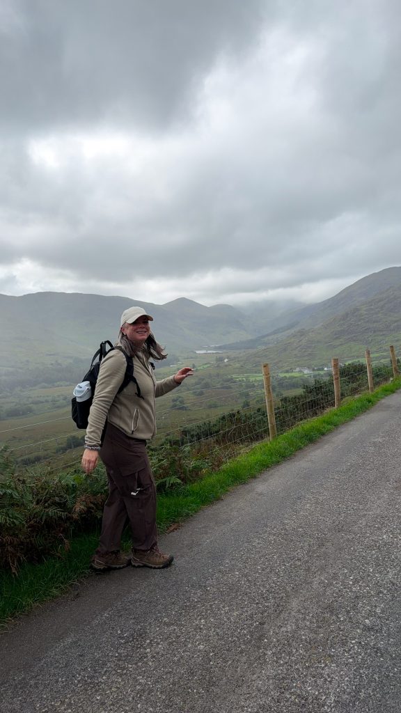 Woman turning towards camera on a paved road with green mountains in the distance on a rainy day.