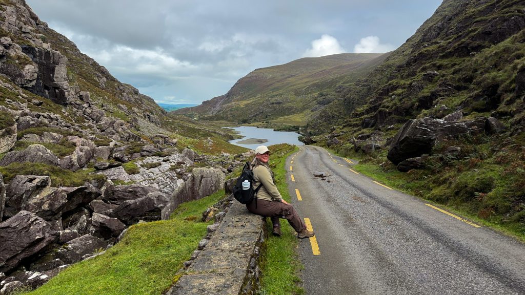 Woman sits on a stone wall next to a paved road that runs between mountains towards a lake in the distance.