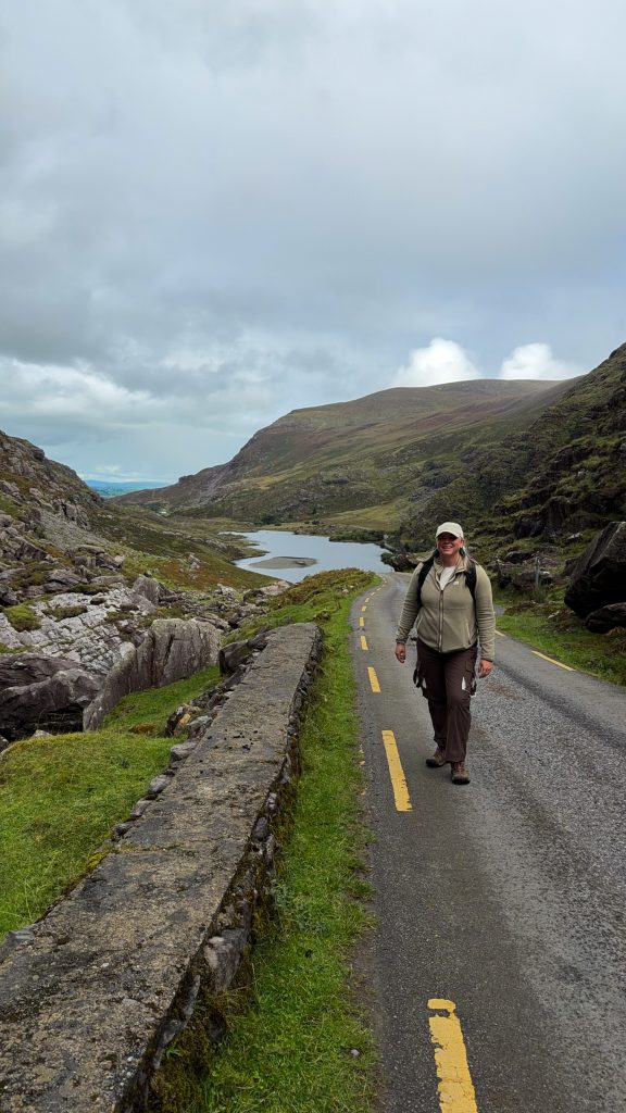 Woman stands on a narrow paved road smiling at the camera with a lake and mountains in the distance.