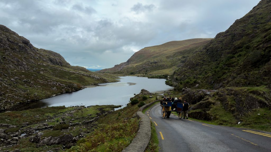 Horse and buggy carries passengers down a narrow road with a lake in the distance surrounded by mountains in the Gap of Dunloe.
