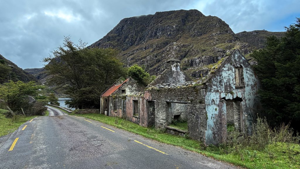 Old cottage in ruins stands next to a paved road with mountains behind it.