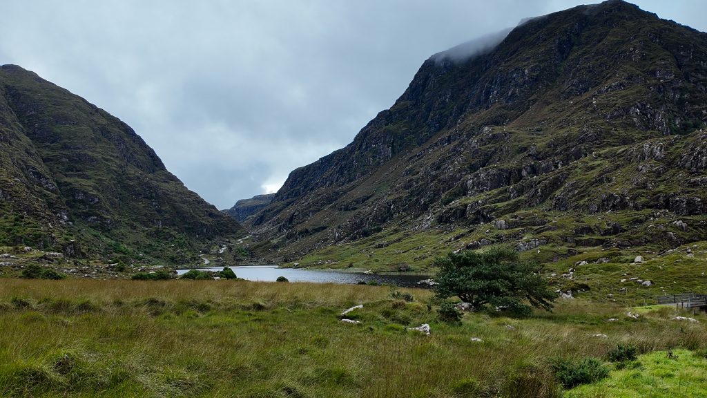 Green fields lead to a small lake beneath two mountain ranges and a narrow road winds between the path.