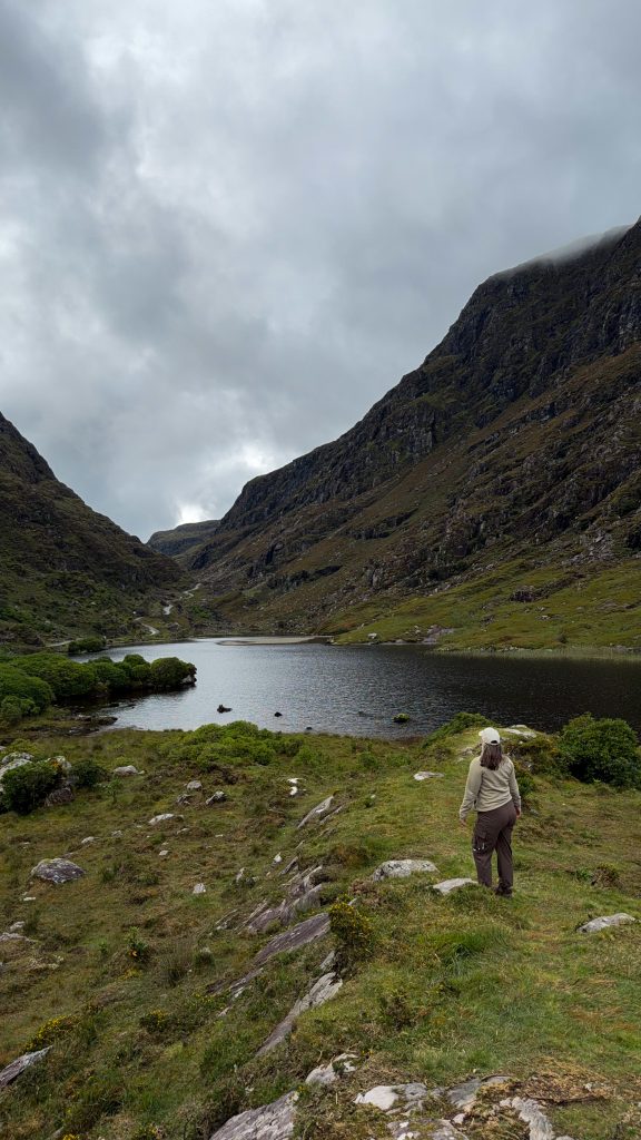 Woman stands overlooking a lake beneath mountain peaks and a windy road leads between the pass in the distance.