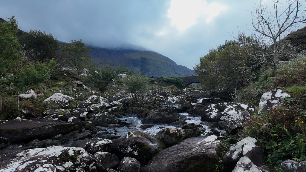 Rocky river leads to an arched stone bridge in the valley of tall mountains on a stormy day.