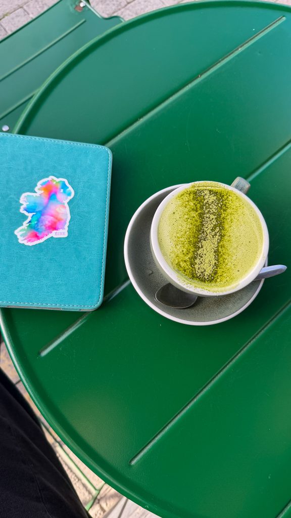 On top of an emerald green table is a green matcha latte in a cup next to a turquoise Kindle with a sticker of Ireland.
