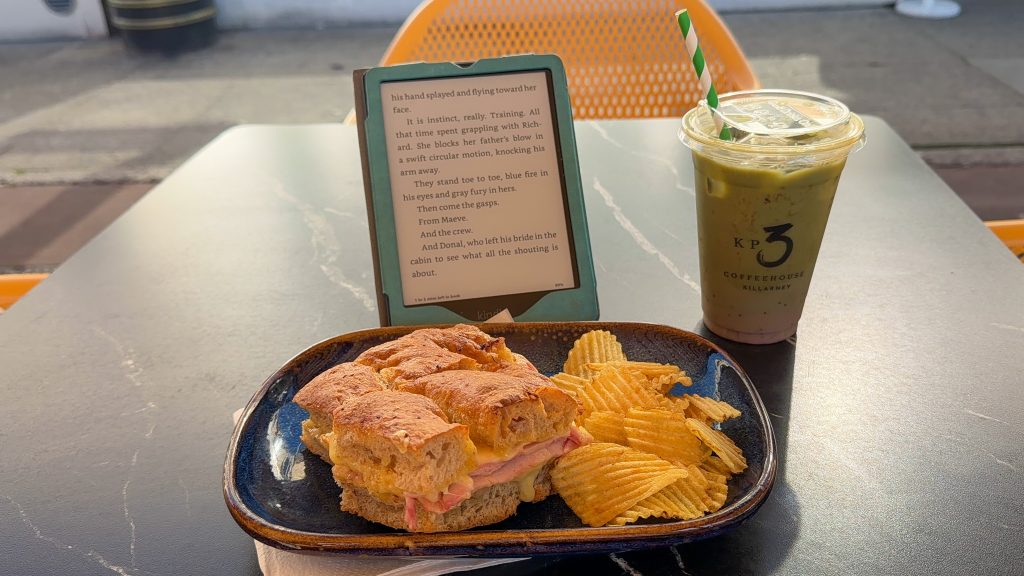 Ham and cheese sandwich next to chips on a plate with a matcha latte and an open Kindle with words on it sit on a table.