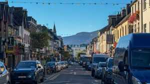 Street in Killarney, Ireland with cars and buildings with mountains and a church spire in the distance. The Best things to do in Killarney, Ireland.