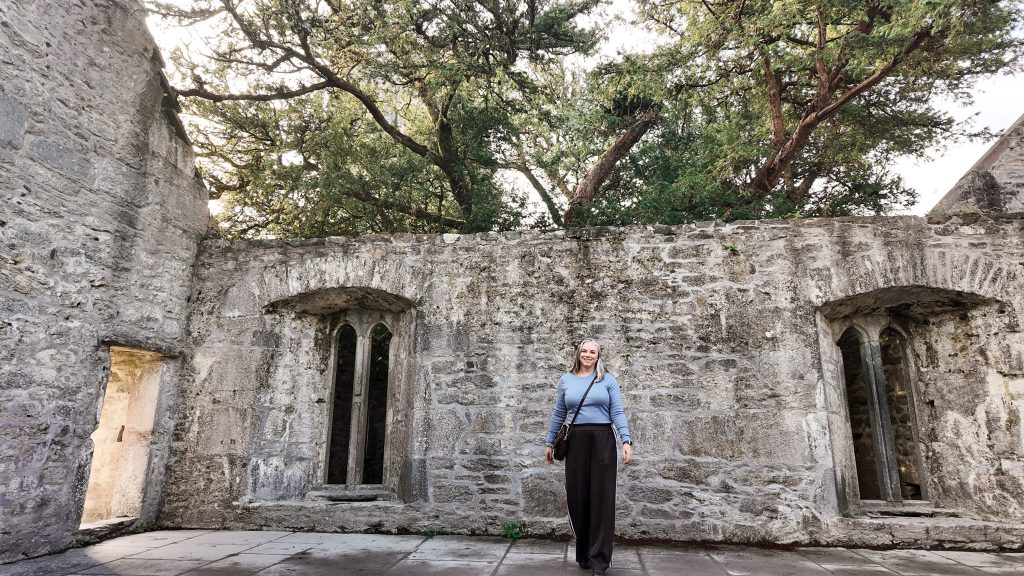 Woman wearing a blue shirt and brown pants stands in front of stone walls with no roof, but the top of a Yew Tree leaves above her.