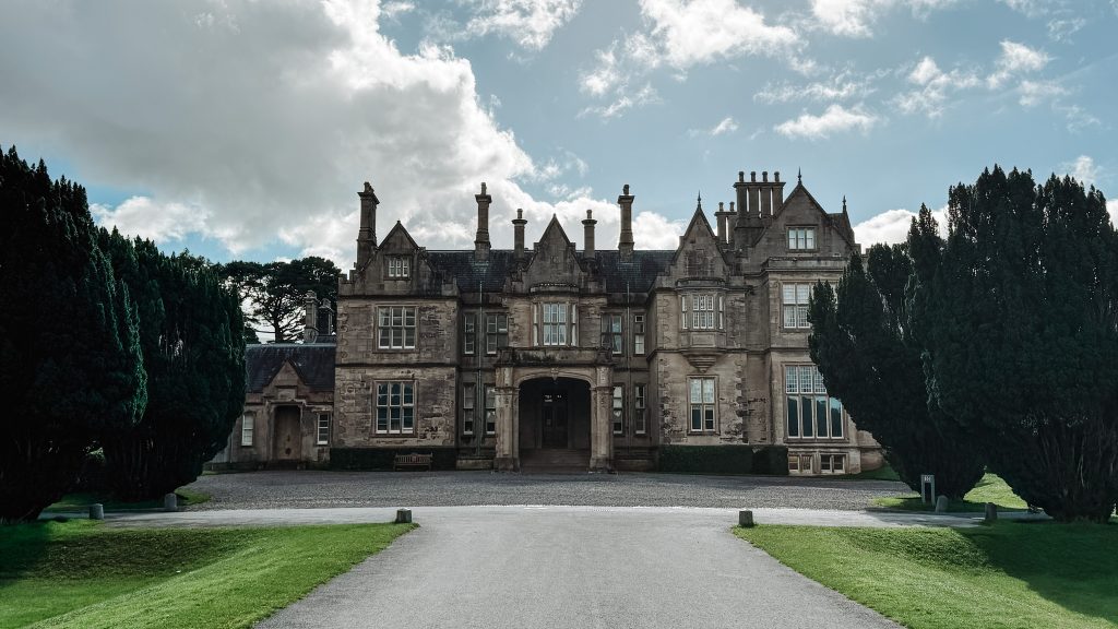 Large tan stone mansion sits at the end of a paved road between two green trees. Muckross House in Killarney National Park.