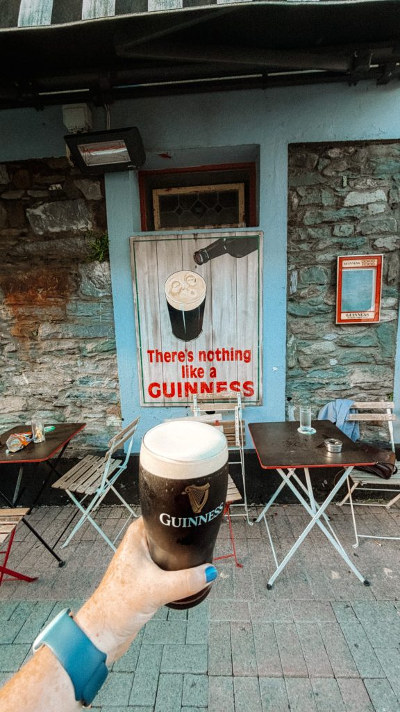 A hand holds a pint of Guinness in front of a stone pub that has a sign about Guinness and folding tables.