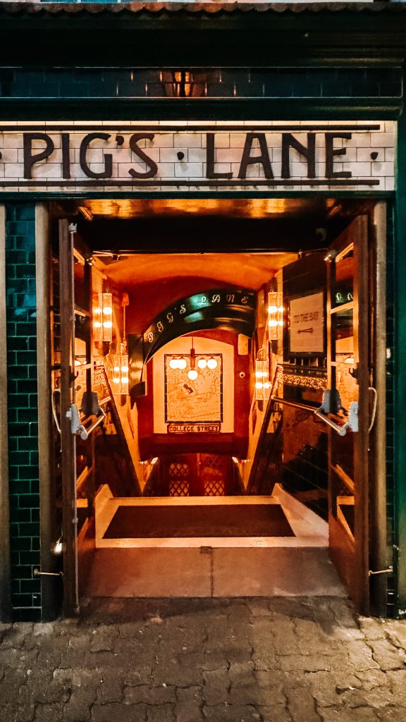 A white tile sign above an entry way reads Pig's Lane and leads to a tiled staircase.