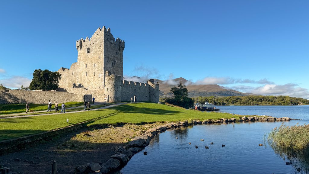 Stone castle sits on green grass next to a lake on a blue sky day. Ross Castle in Killarney National Park.