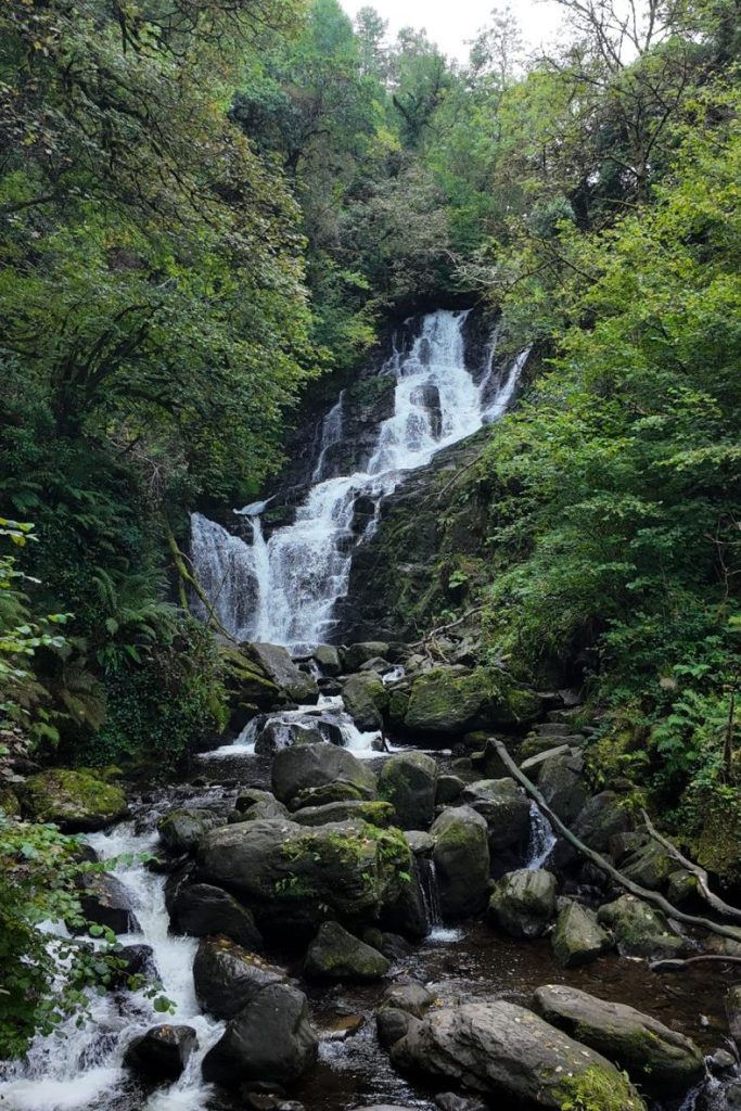 Tall waterfall flows over rocks in between green trees.